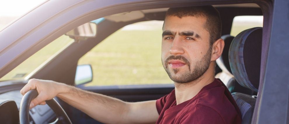 Um homem de camiseta vermelha está sentado dentro de seu carro, com a mão direita sobre o volante. Ele mira o observador, mostrando seu rosto completo. O homem tem barba cerrada, cabelos curtos e um traço largo feito a navalha na sobrancelha.