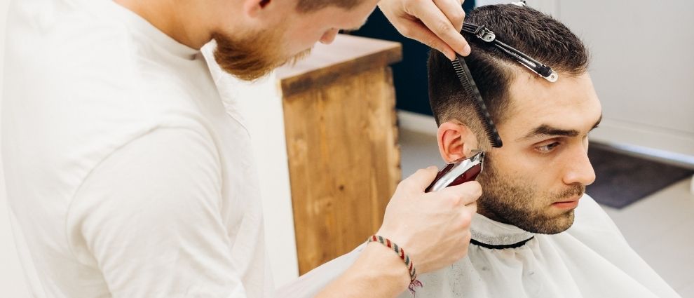 Um homem de semblante sério está sentado na cadeira de uma barbearia, usando um avental branco. Ele tem os cabelos presos com pinças, para facilitar o trabalho do barbeiro, que está de pé com uma máquina, aparando as costeletas do cliente.