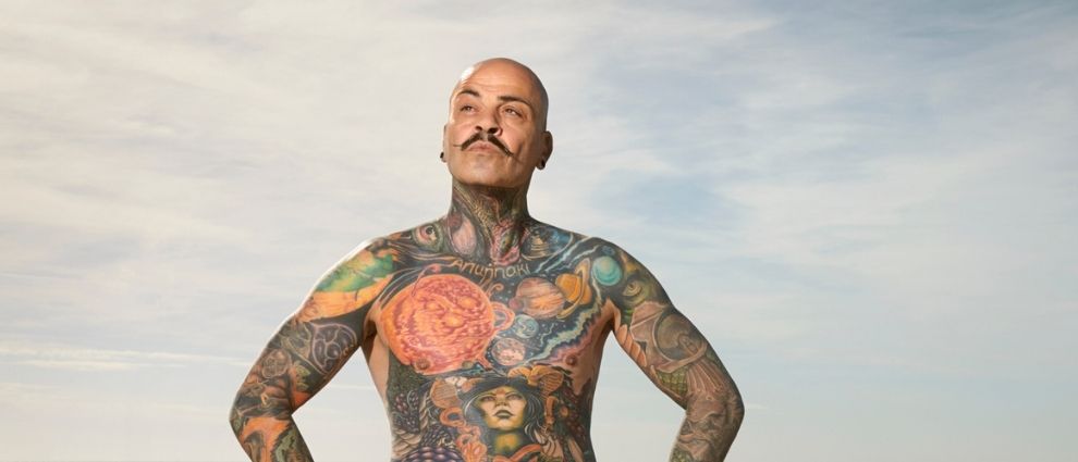 Homem tatuado sem camisa na praia.