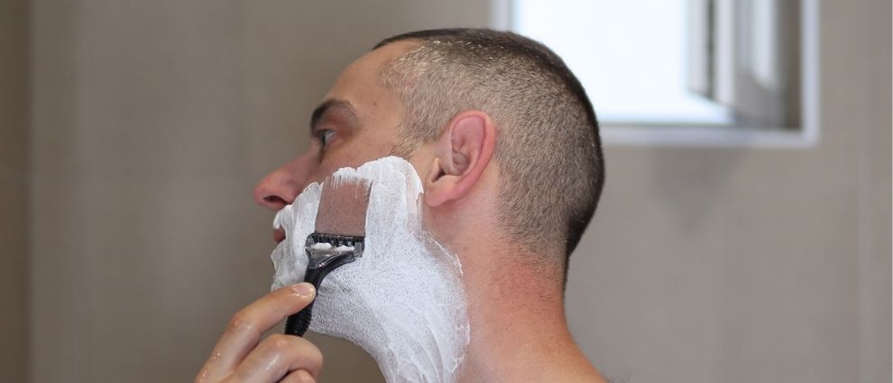 Homem com espuma de barbear no rosto raspando os pelos da face com uma lâmina de babear.