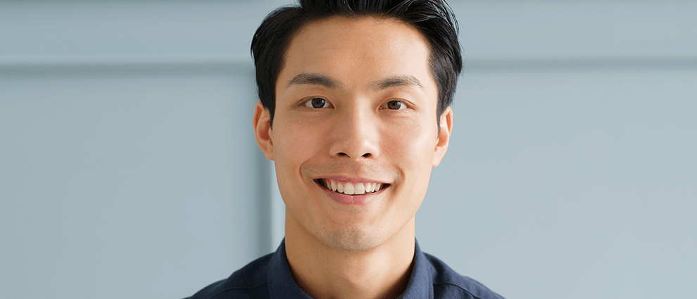 Homem asiático adulto, com camisa social preta, sem barca e com corte de cabelo side swept, sorri em foto frontal.
